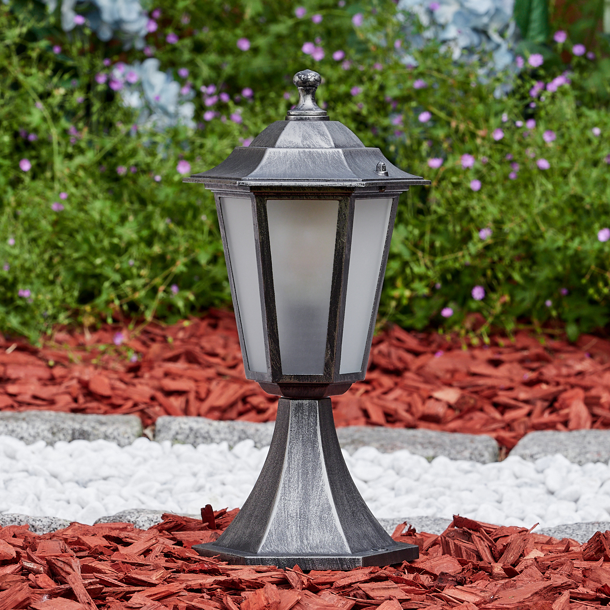 Eine silberne Außenlampe steht auf roter Mulch, weißen Steinen und einer Steinumrandung. Grünes Laub und lila Blumen sind im Hintergrund.