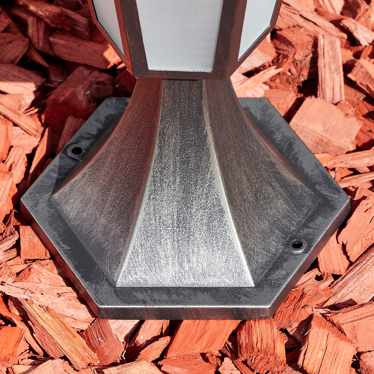 Ein Metallsockel steht auf roter Mulch. Die Lampe hat eine dekorative Oberseite mit Glas. Der Sockel ist sechseckig.