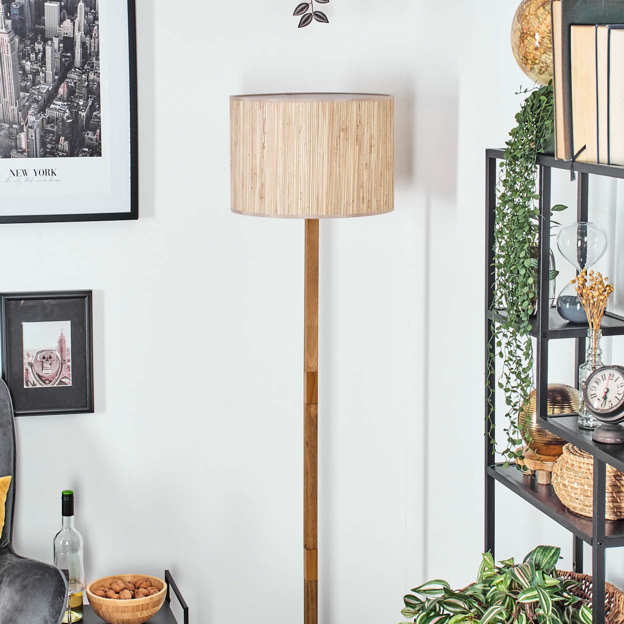Ein Raum mit einer hölzernen Stehlampe und Bildern an der weißen Wand. Ein Regal mit Büchern und Pflanzen befindet sich rechts.