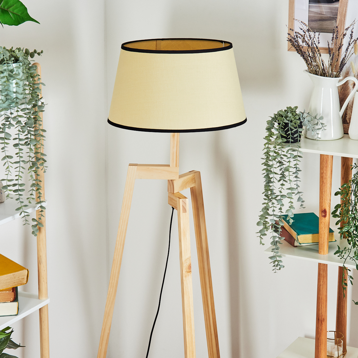 Eine Stehlampe mit cremefarbenem Schirm und Holzbeinen steht in einem Raum mit Pflanzen und Regalen.