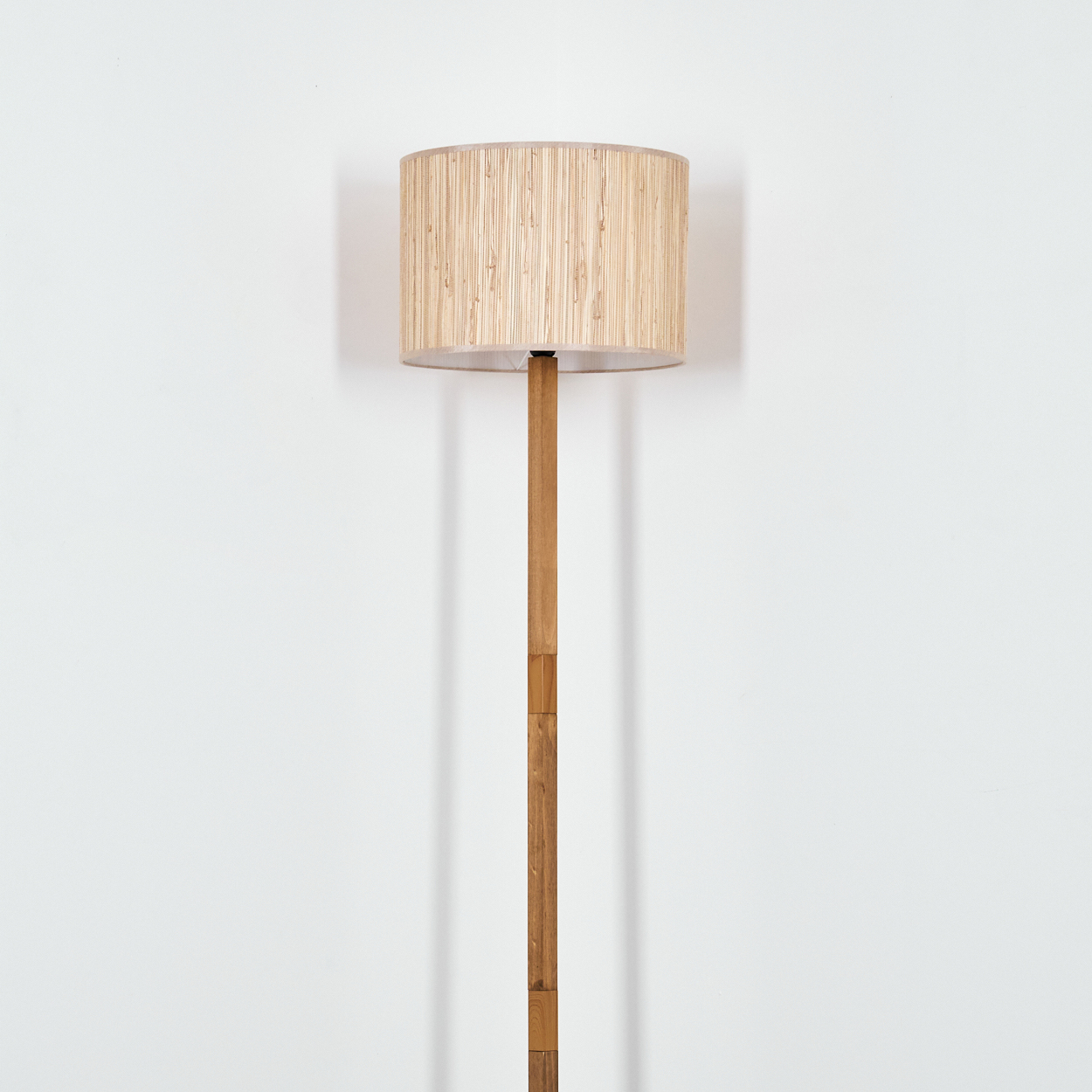 Eine hohe Stehlampe aus Bambus mit zylindrischem Schirm steht vor einer weißen Wand. Der Schirm ist beige und die Stange braun.
