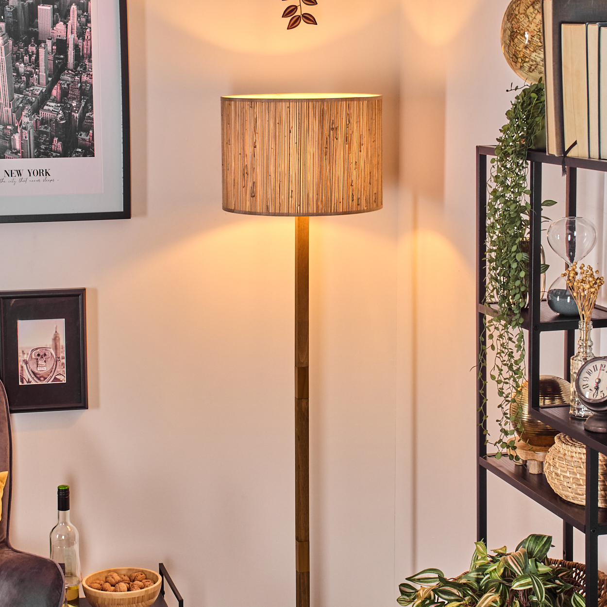 Eine Stehlampe mit Holzfuß und Lampenschirm. Rechts ein Regal mit Pflanzen, Büchern und einem Globus. Ein Bild an der Wand.