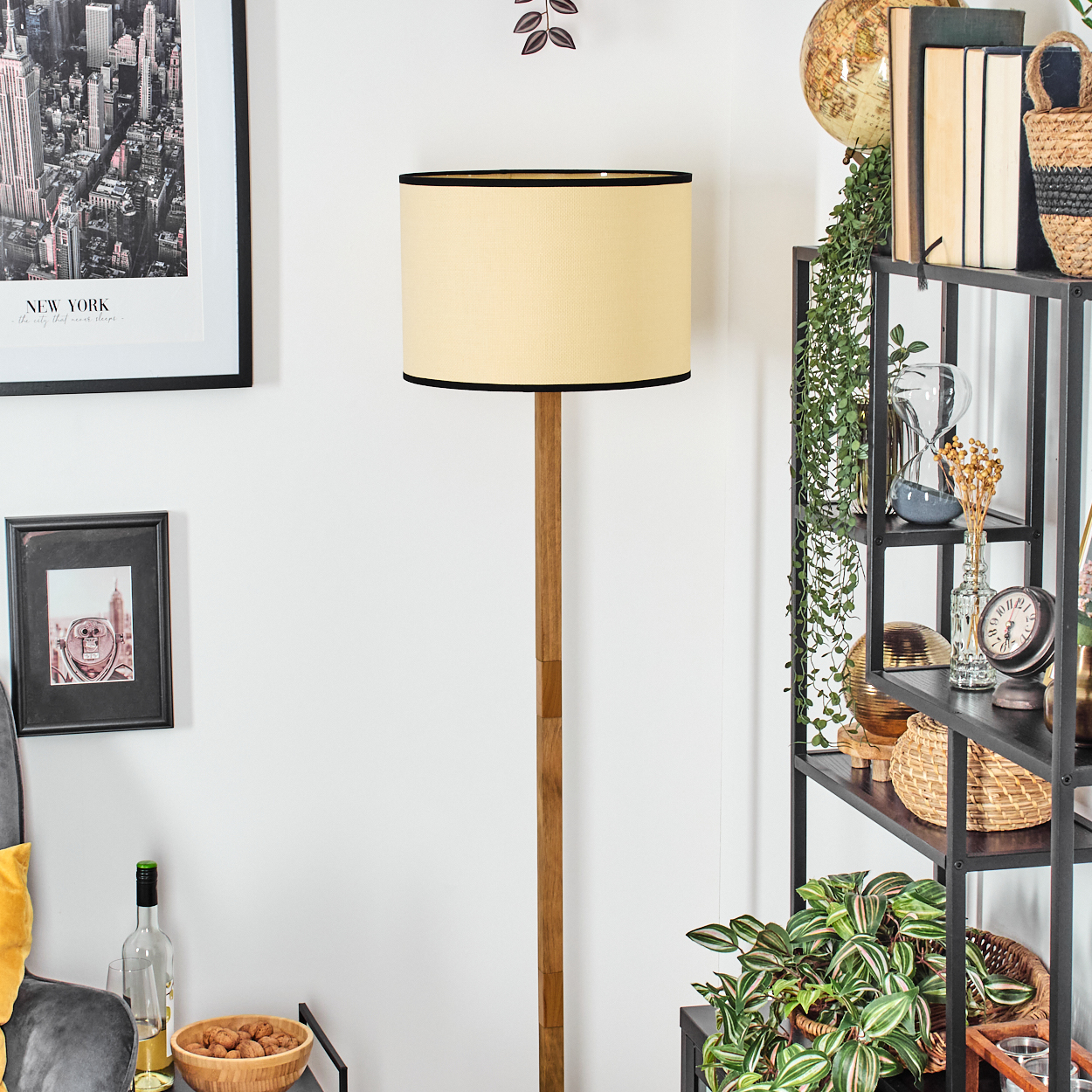 Eine Stehlampe mit beigefarbenem Schirm steht neben einem schwarzen Regal mit Dekorationsartikeln und Pflanzen. Bilder hängen an der Wand.