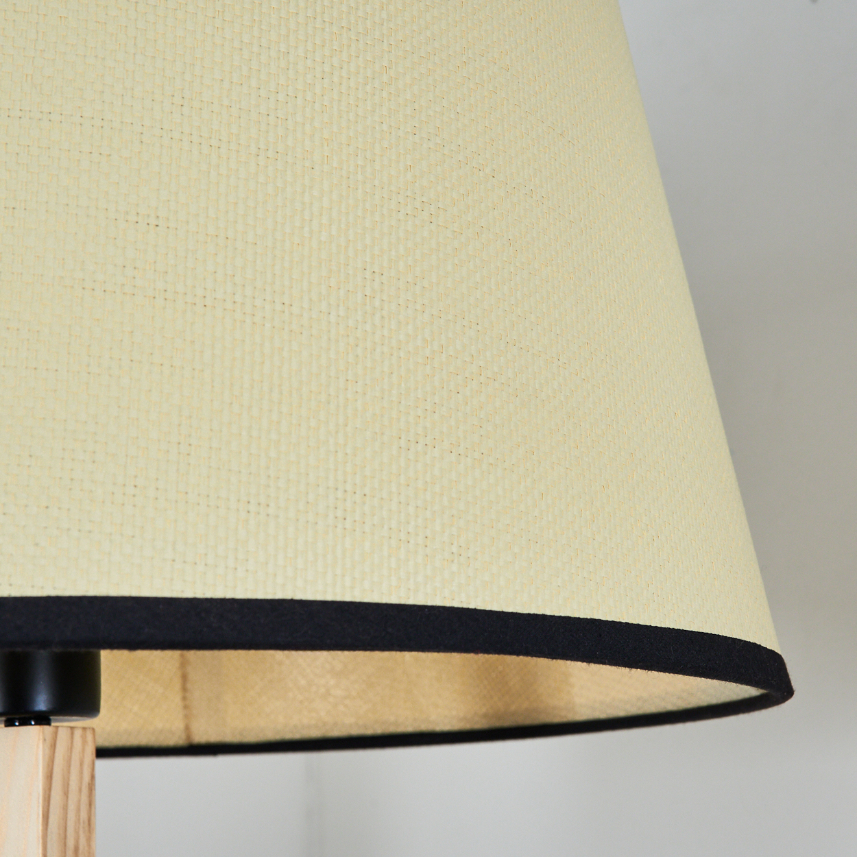 Nahaufnahme einer Lampe. Der Lampenschirm ist cremefarben mit schwarzem Besatz. Der Sockel ist aus Holz und schwarz. Der Hintergrund ist weiß.