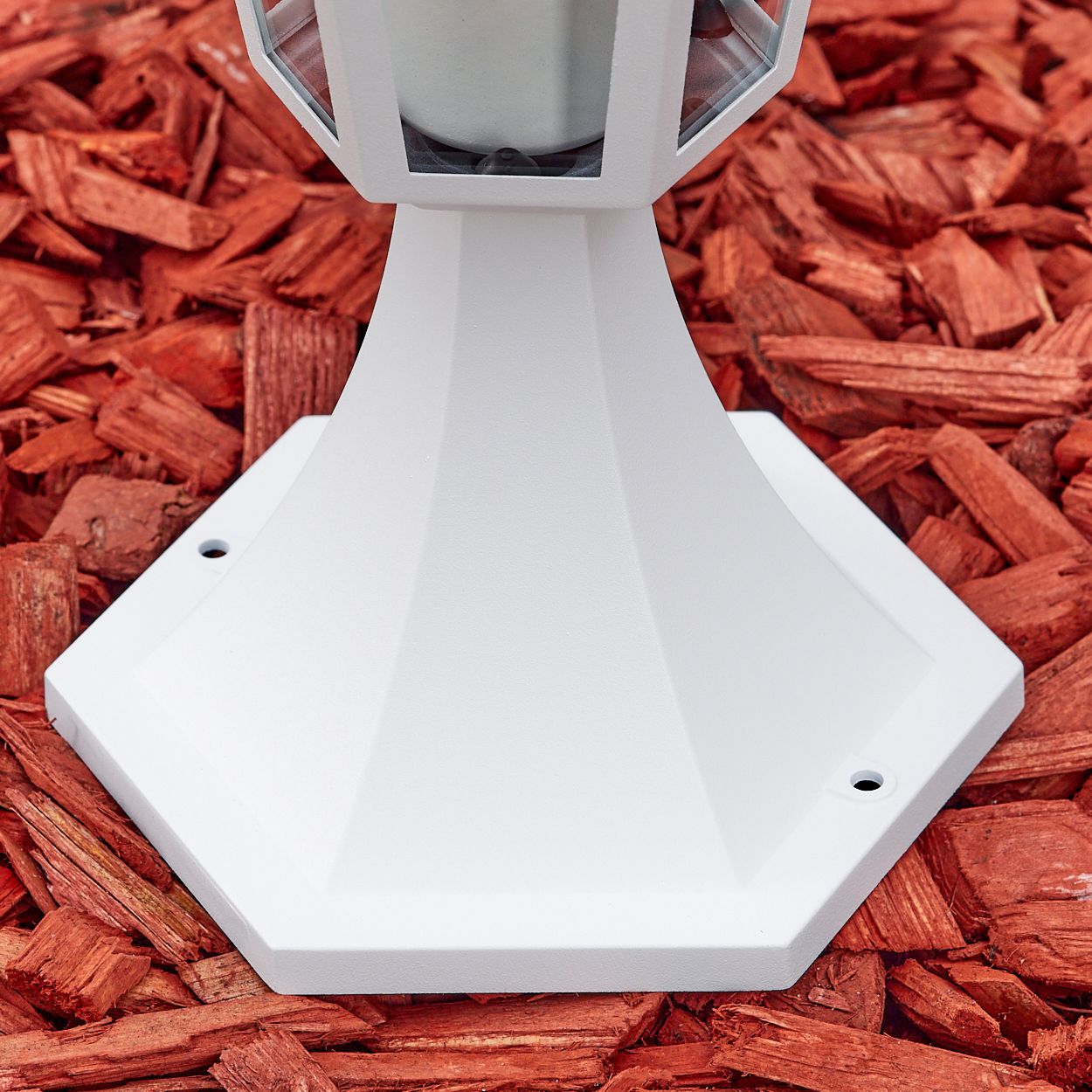 Ein weißer Lampenfuß auf roter Mulch. Die Basis ist achteckig und verjüngt sich zu einer Lampe mit Glasscheiben.