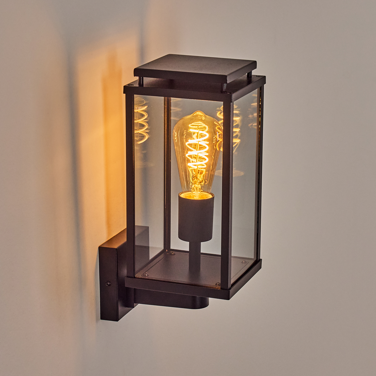 Eine schwarze Wandlaterne mit Glasscheiben, in der sich eine leuchtende Glühbirne mit Spiralfaden befindet. An einer weißen Wand montiert.