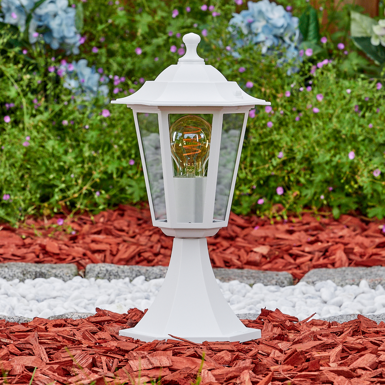 Weiße Außenlampe mit brennender Glühbirne. In einem Garten mit roter Mulch, weißen Steinen und grünem Laub mit blauen und lila Blüten.