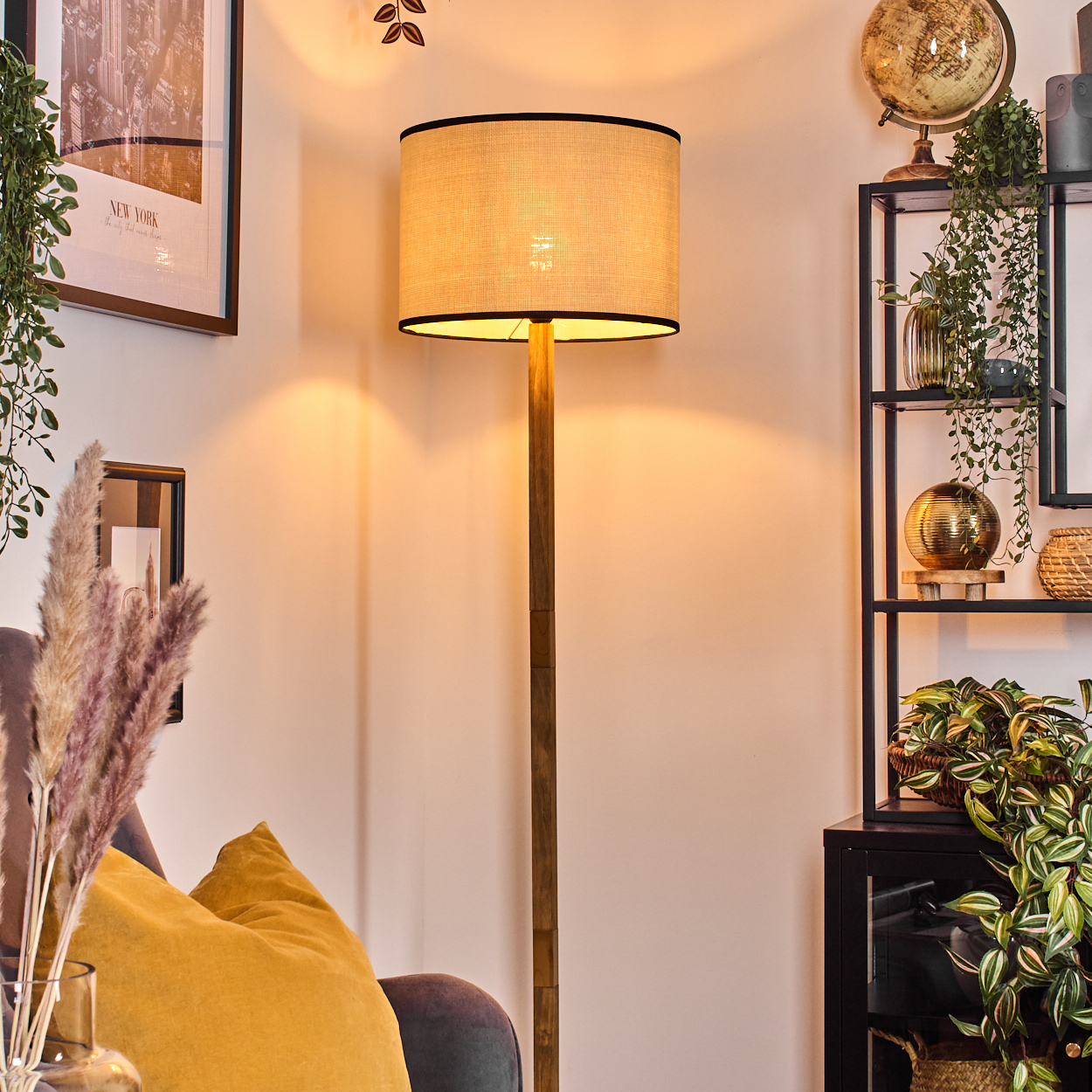 Eine Stehlampe mit beigem Schirm steht in einem gemütlichen Wohnzimmer. Pflanzen und Dekoration schaffen eine warme Atmosphäre.