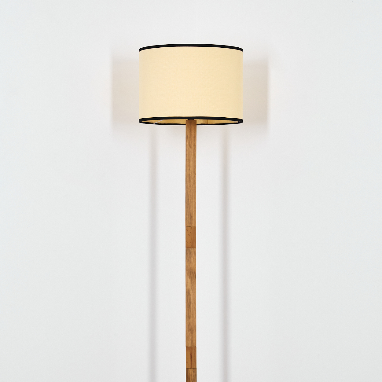 Eine hohe Stehlampe aus Holz mit beigem Lampenschirm und schwarzem Rand steht vor einer weißen Wand. Die Lampe ist zentriert.