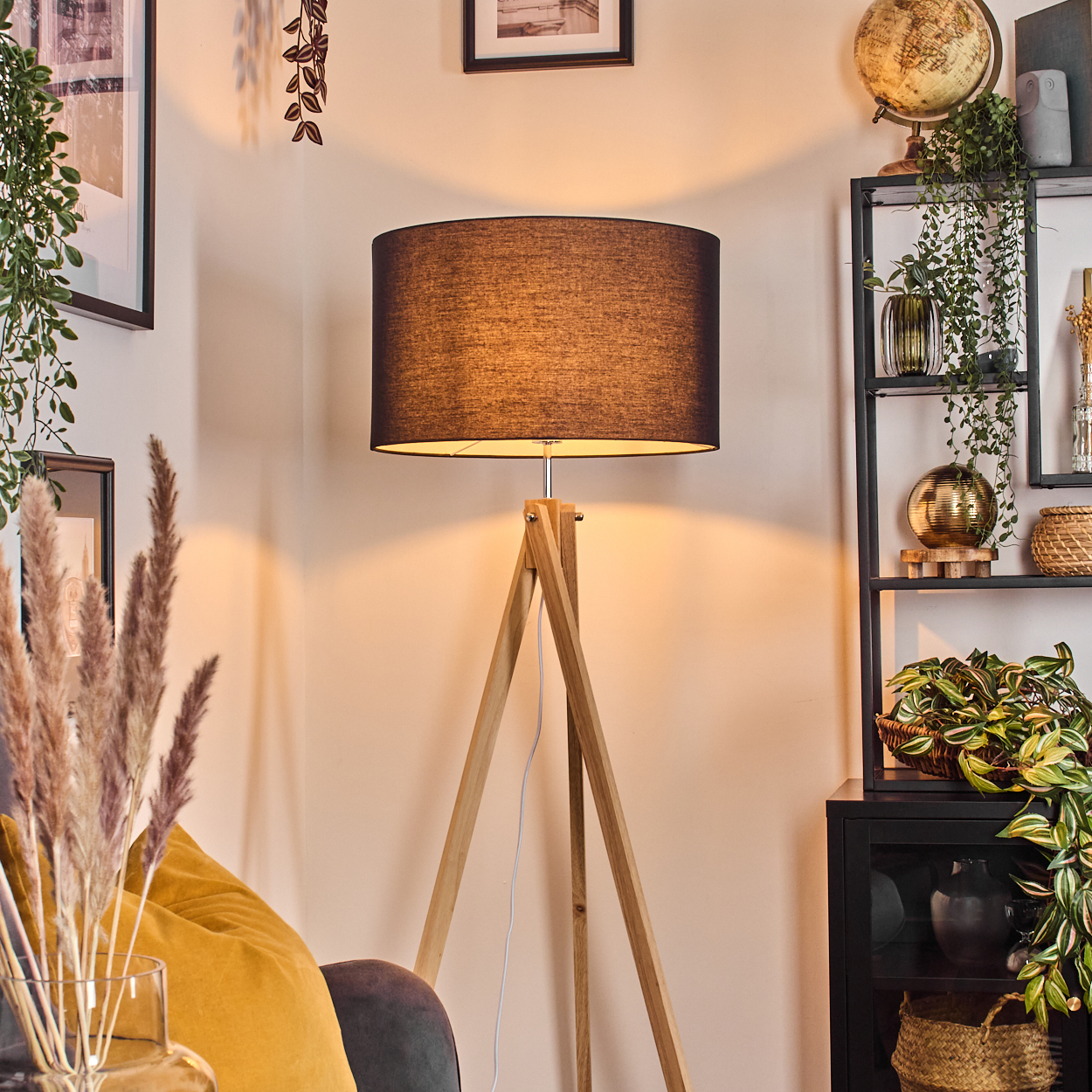 Eine Stehlampe mit schwarzem Lampenschirm steht in einem Raum. Es gibt Pflanzen, ein gelbes Kissen und ein Regal mit Dekoration.