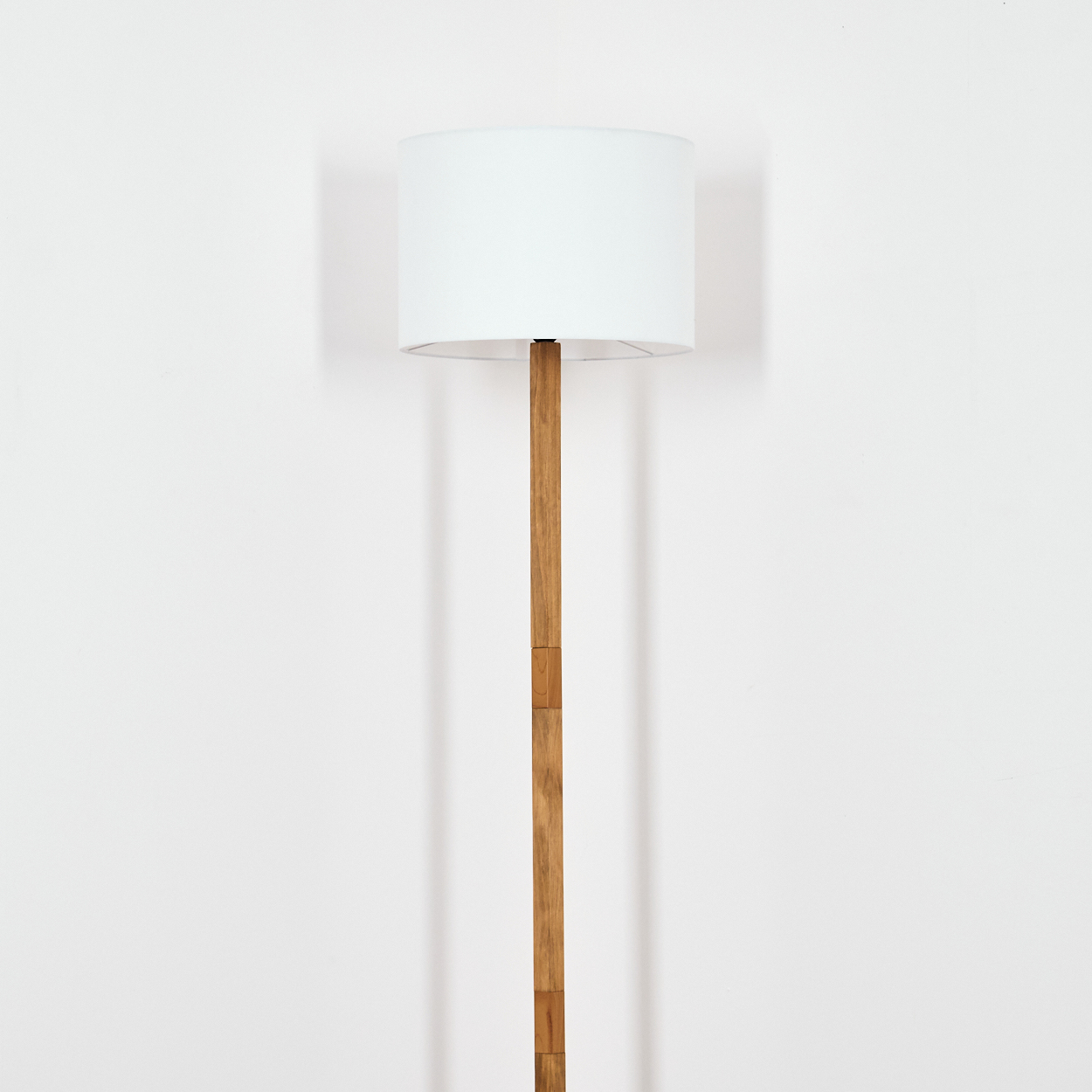 Eine Stehlampe steht vor einer weißen Wand. Sie hat einen Holzständer und einen weißen Lampenschirm. Die Lampe wirft einen Schatten.