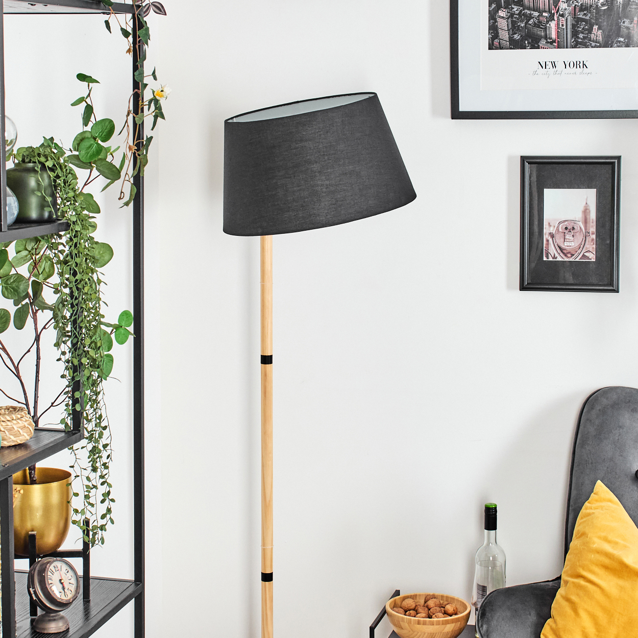 Eine schwarze Lampe mit Holzfuß steht neben einer Wand mit Kunst. Ein Stuhl mit gelbem Kissen steht daneben. Pflanzen und ein Regal sind sichtbar.