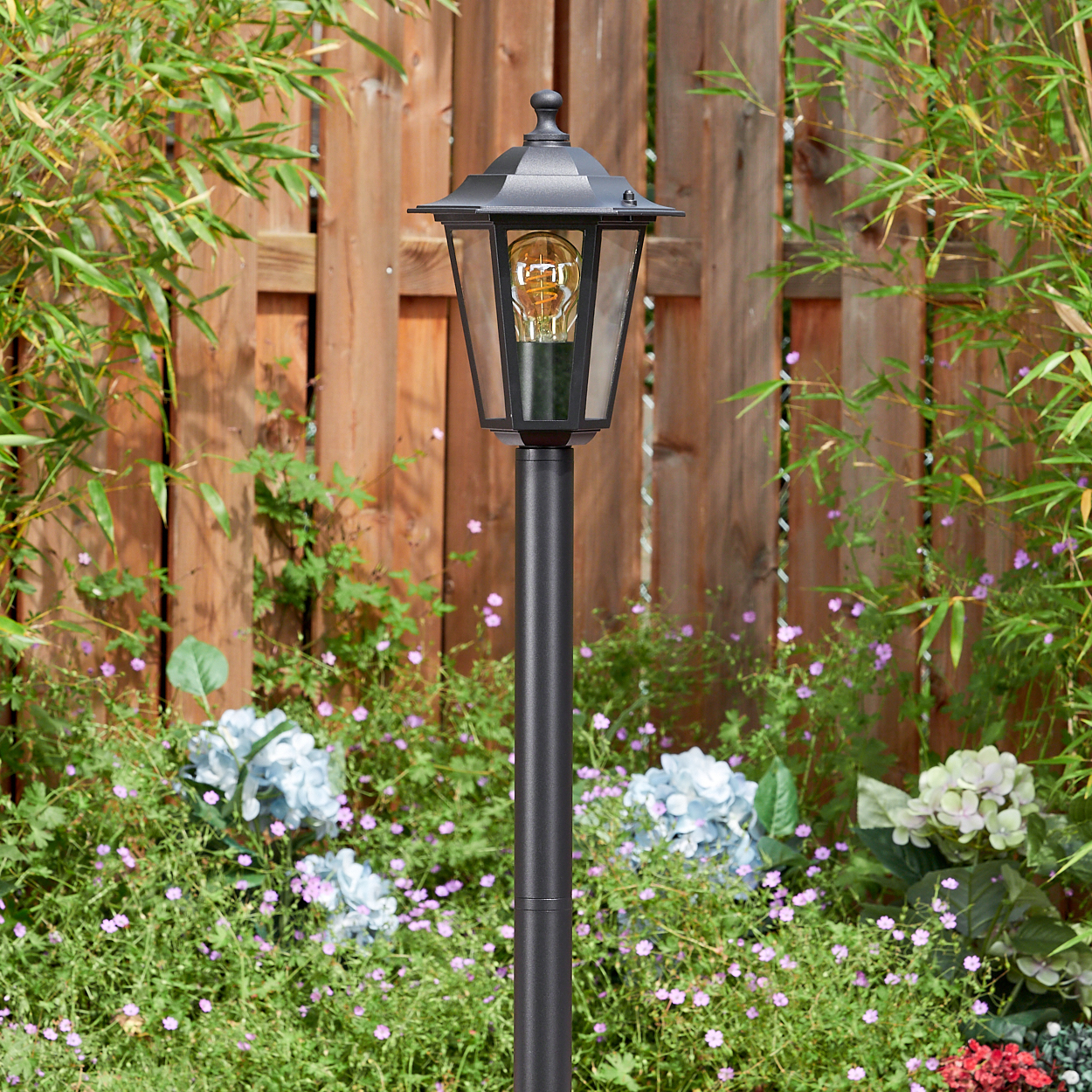 Eine schwarze Außenlampe steht in einem Garten. Im Hintergrund befinden sich ein Holzzaun und Grün mit blauen und lila Blumen.