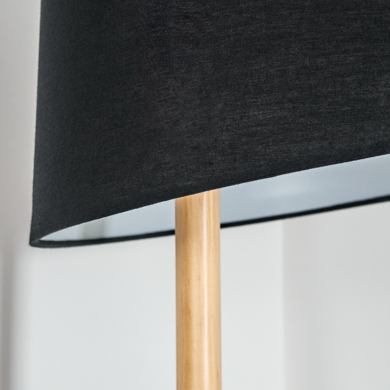 Nahaufnahme einer Lampe. Sie hat einen schwarzen Schirm und eine helle Holzstange. Der Hintergrund ist eine weiße Wand.