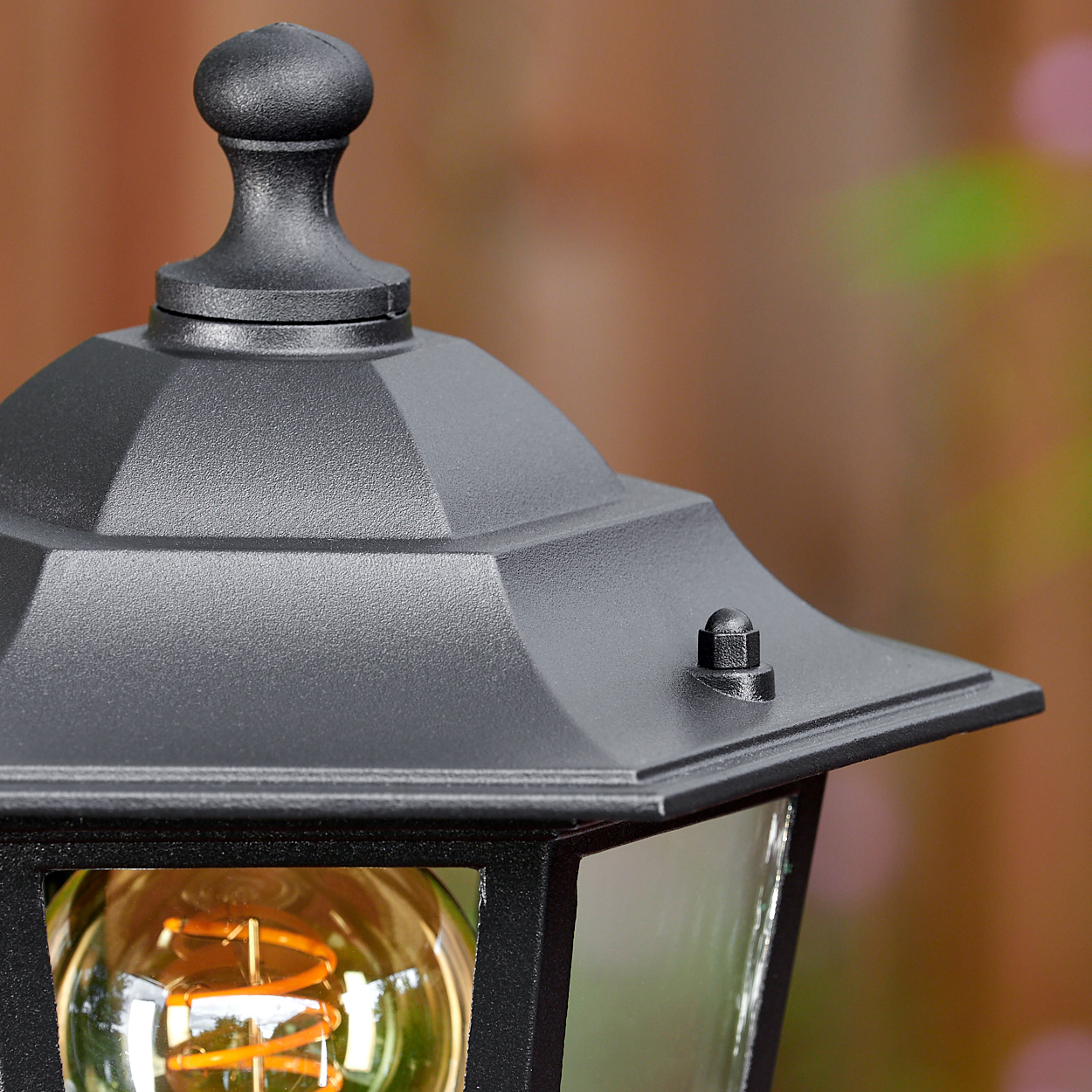 Nahaufnahme einer schwarzen Außenlampe. Die Lampe hat eine Glasscheibe und eine Wendelglühbirne. Der Hintergrund ist verschwommen grün und braun.