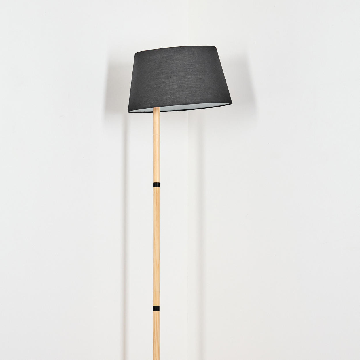 Eine Stehlampe mit schwarzem Lampenschirm und Holzstange steht vor einer weißen Wand. Die Lampe ist abgewinkelt.