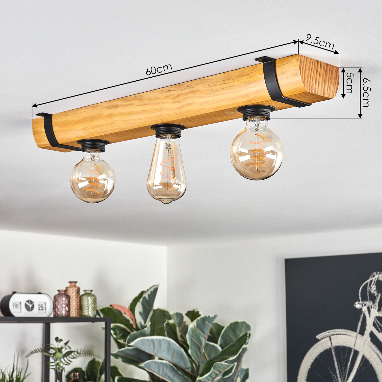 Eine Deckenleuchte aus Holz mit drei Edison-Glühbirnen. Schwarze Metallstützen und die Abmessungen werden angezeigt. Der Hintergrund zeigt Pflanzen und Wandkunst.