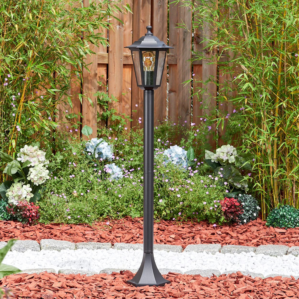 Eine schwarze Straßenlaterne in einem Garten. Dahinter befinden sich ein Holzzaun, Bambus und Blumen. Der Garten hat roten Mulch und weiße Steine.