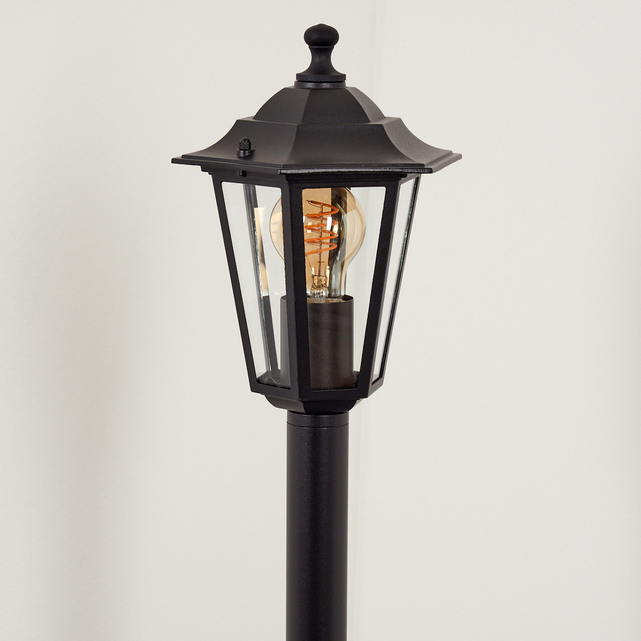 Eine schwarze Außenlampe mit Glasgehäuse, sichtbarer Glühbirne und dekorativer Spitze, vor einer weißen Wand.