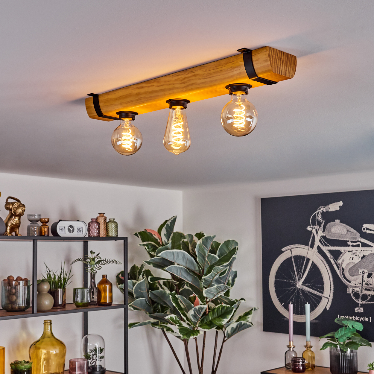 Holzbalken-Deckenleuchte mit drei Glühbirnen. Darunter eine Pflanze, ein Regal mit Dekoration und ein Motorrad-Kunstwerk.