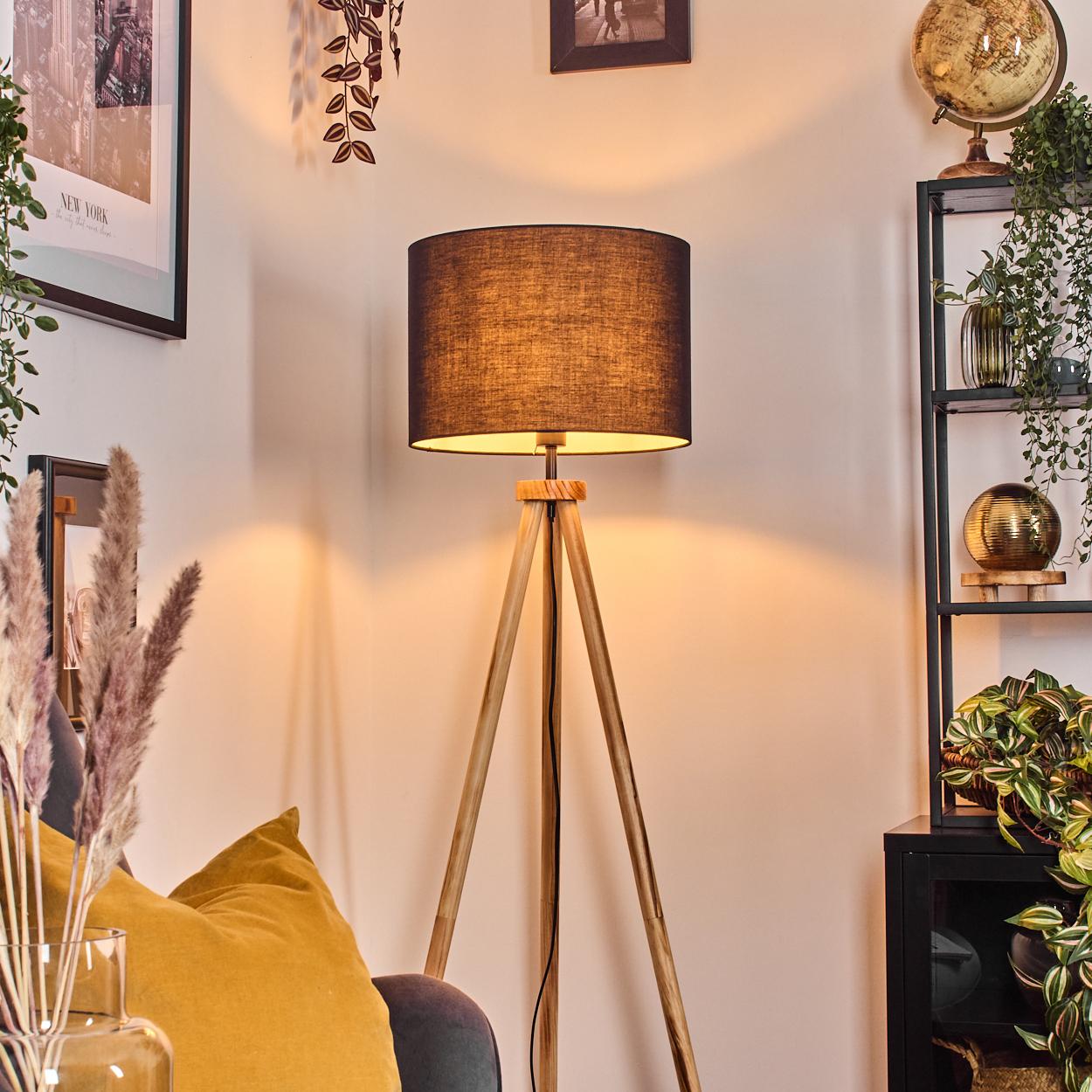 Eine hölzerne Stativ-Stehlampe mit schwarzem Lampenschirm steht in der Nähe eines Sofas mit gelben Kissen, einem Regal mit Dekoration und einem Globus.