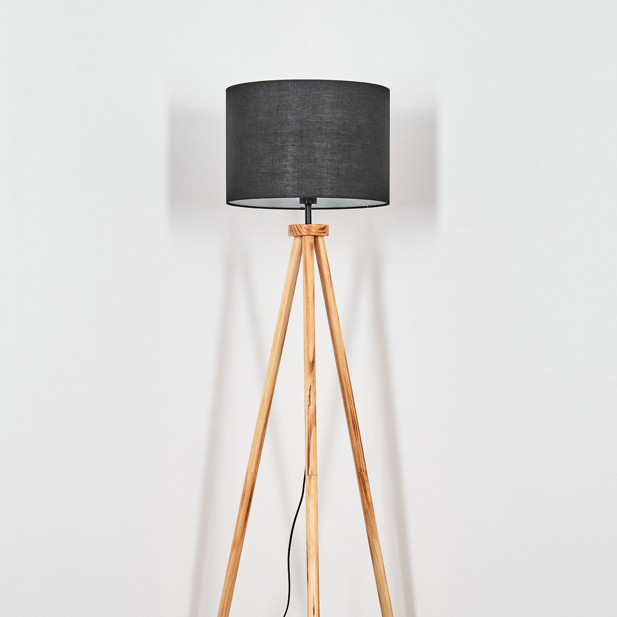 Eine Stehlampe mit schwarzem Lampenschirm und Holzstativfuß steht vor einer weißen Wand. Ein schwarzes Kabel verläuft die Beine hinunter.