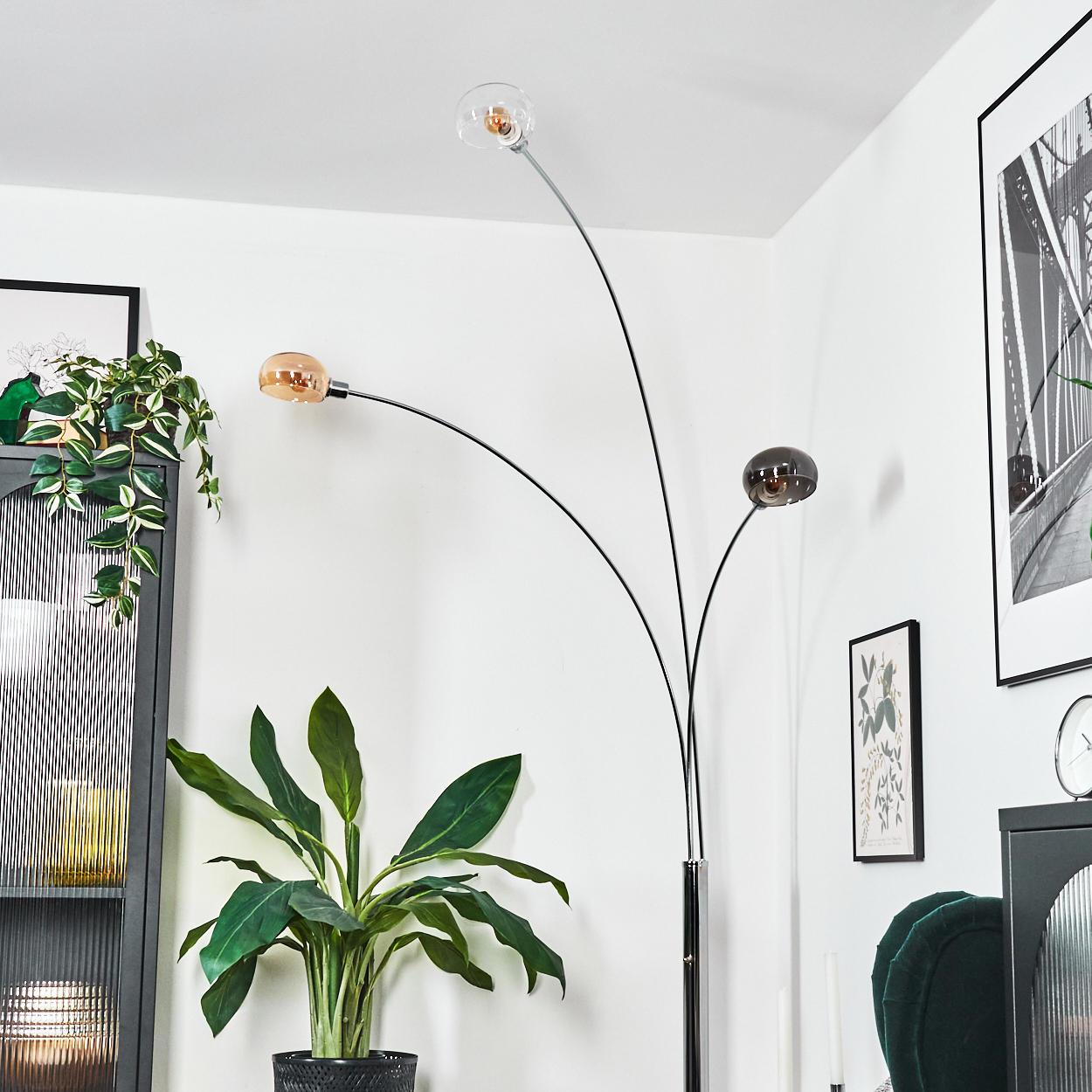 Eine moderne Stehlampe mit drei Glasschirmen. Eine große grüne Pflanze in einem schwarzen Topf ist im Vordergrund. Kunstwerke und Möbel sind im Hintergrund.