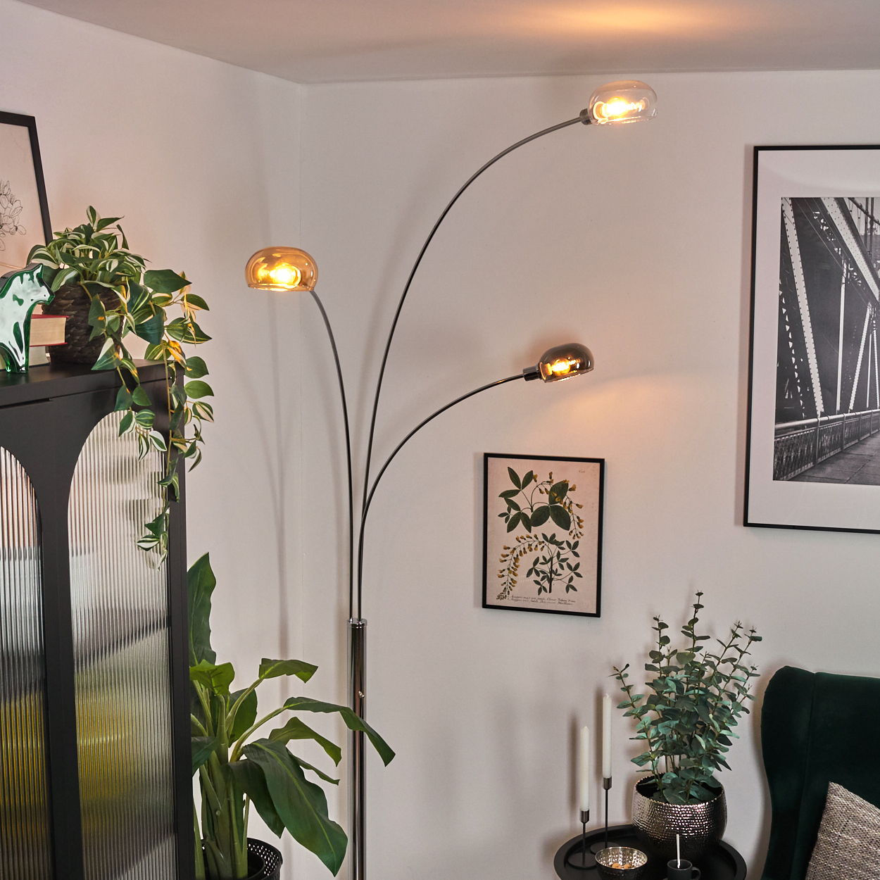 Eine moderne Stehlampe mit drei Lichtern beleuchtet einen Raum mit Pflanzen, einem schwarzen Schrank und gerahmten Kunstwerken.