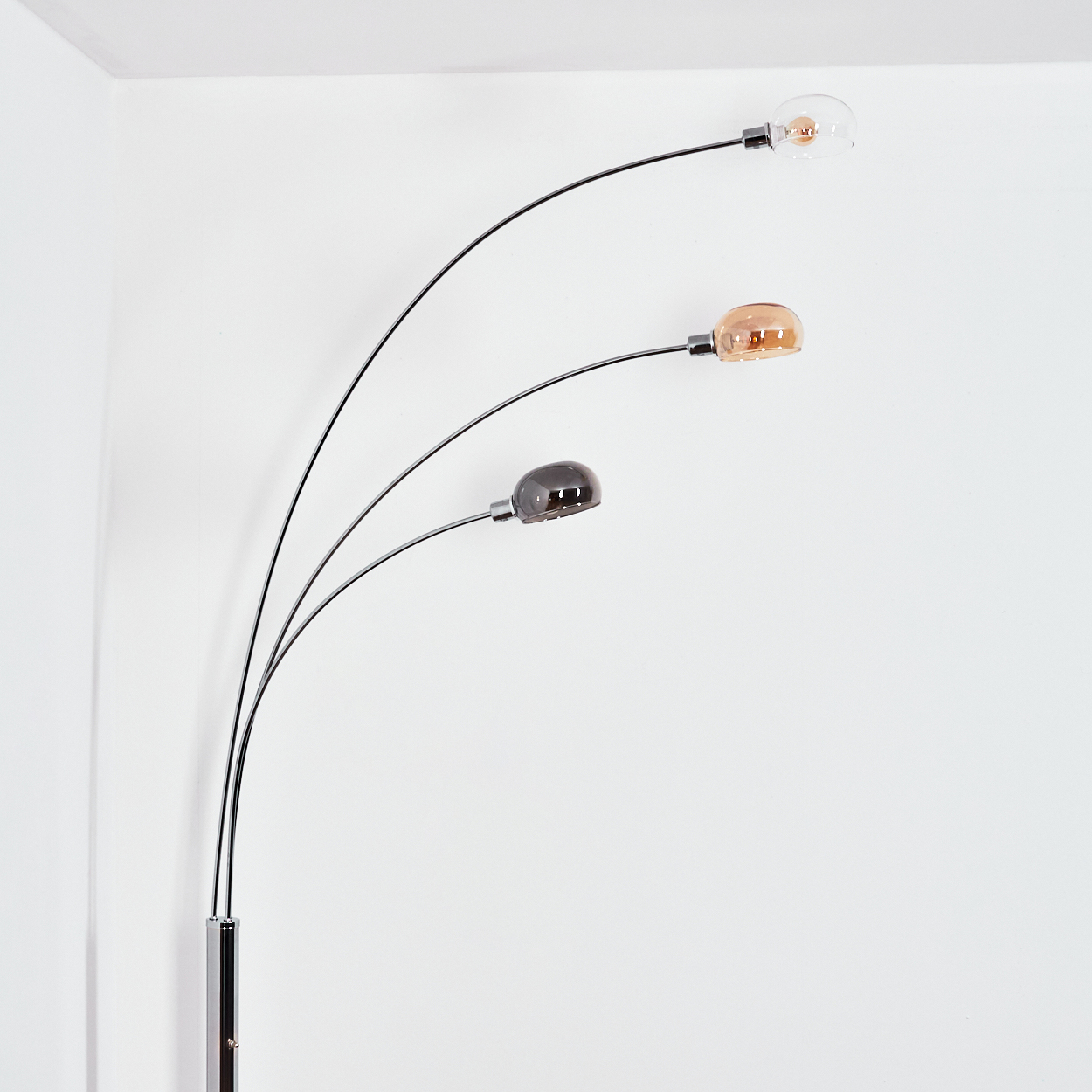 Eine moderne Stehlampe mit drei gebogenen Armen. Jeder Arm hält eine Glühbirne in verschiedenen Farben: klar, bernsteinfarben und dunkelgrau, vor einer weißen Wand.
