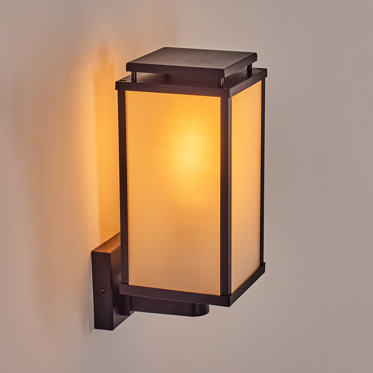 Eine rechteckige Wandleuchte mit bronzefarbenem Rahmen. Die Lampe strahlt ein warmes, diffuses Licht aus. Sie ist an einer hellbeigen Wand befestigt.
