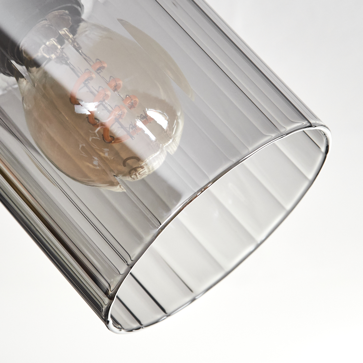 Nahaufnahme einer Leuchte. Ein klarer, gerippter Glasschirm beherbergt eine Glühbirne mit einem orangefarbenen Glühfaden. Der Hintergrund ist weiß.