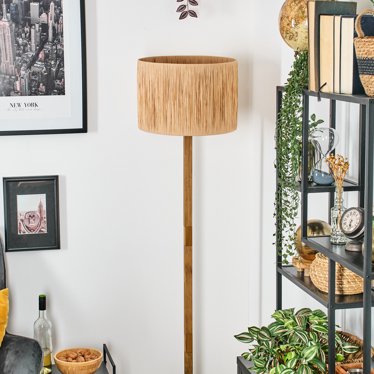 Eine Stehlampe mit gewebtem Lampenschirm steht neben einem Regal mit Dekoration. Bilder hängen an der weißen Wand. Eine Schüssel mit Nüssen steht in der Nähe.