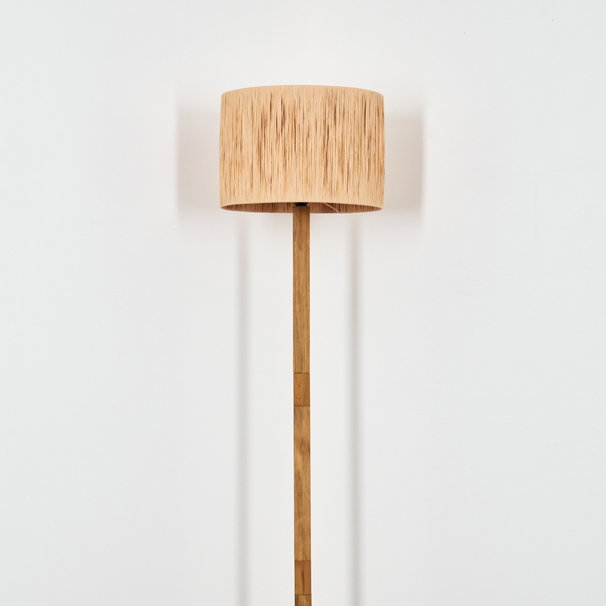 Eine Stehlampe steht vor einer weißen Wand. Sie hat einen beigefarbenen Lampenschirm und eine braune Holzstange. Der Hintergrund ist weiß.
