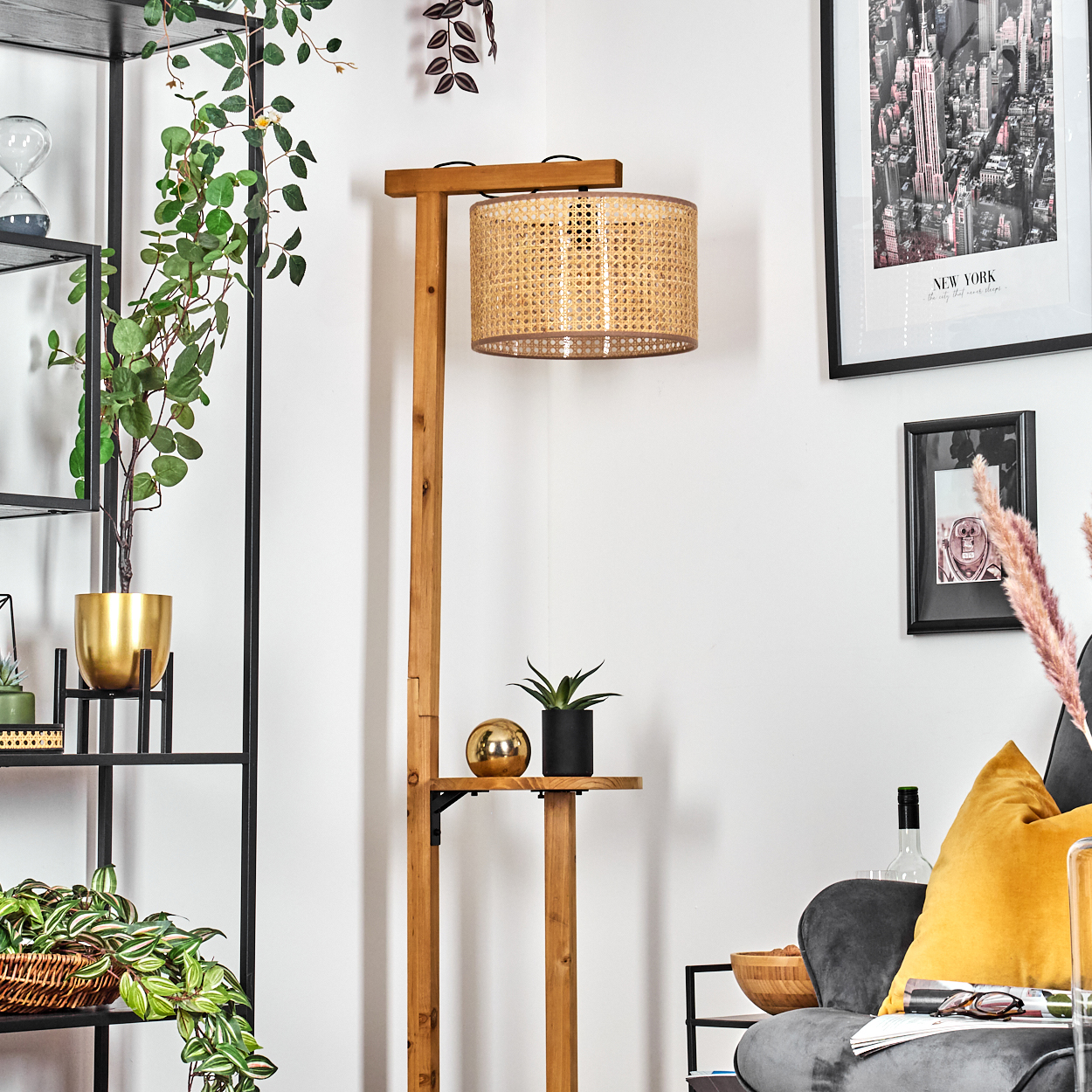 Eine Holz-Stehlampe mit gewebtem Lampenschirm. Ein Regal hält eine Pflanze und eine goldene Kugel. Pflanzen und gerahmte Kunst schmücken die weiße Wand.