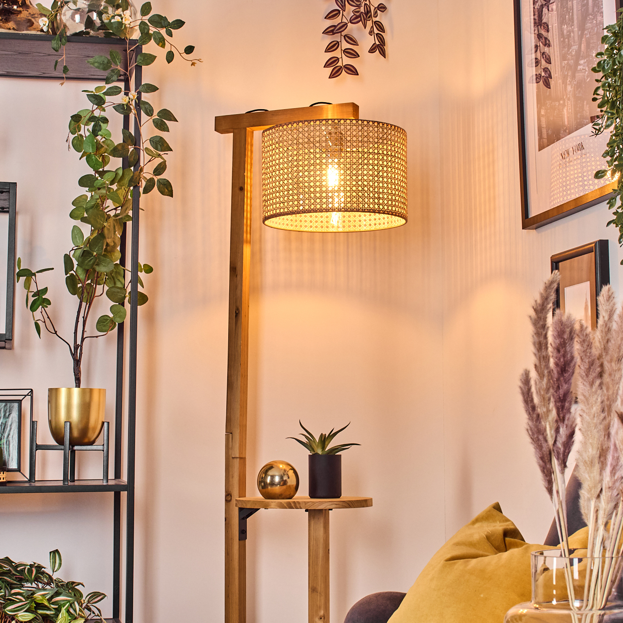 Eine Holzstehlampe mit gewebtem Lampenschirm ist beleuchtet. Auf einem Regal befinden sich Pflanzen, eine goldene Kugel und ein schwarzer Blumentopf.