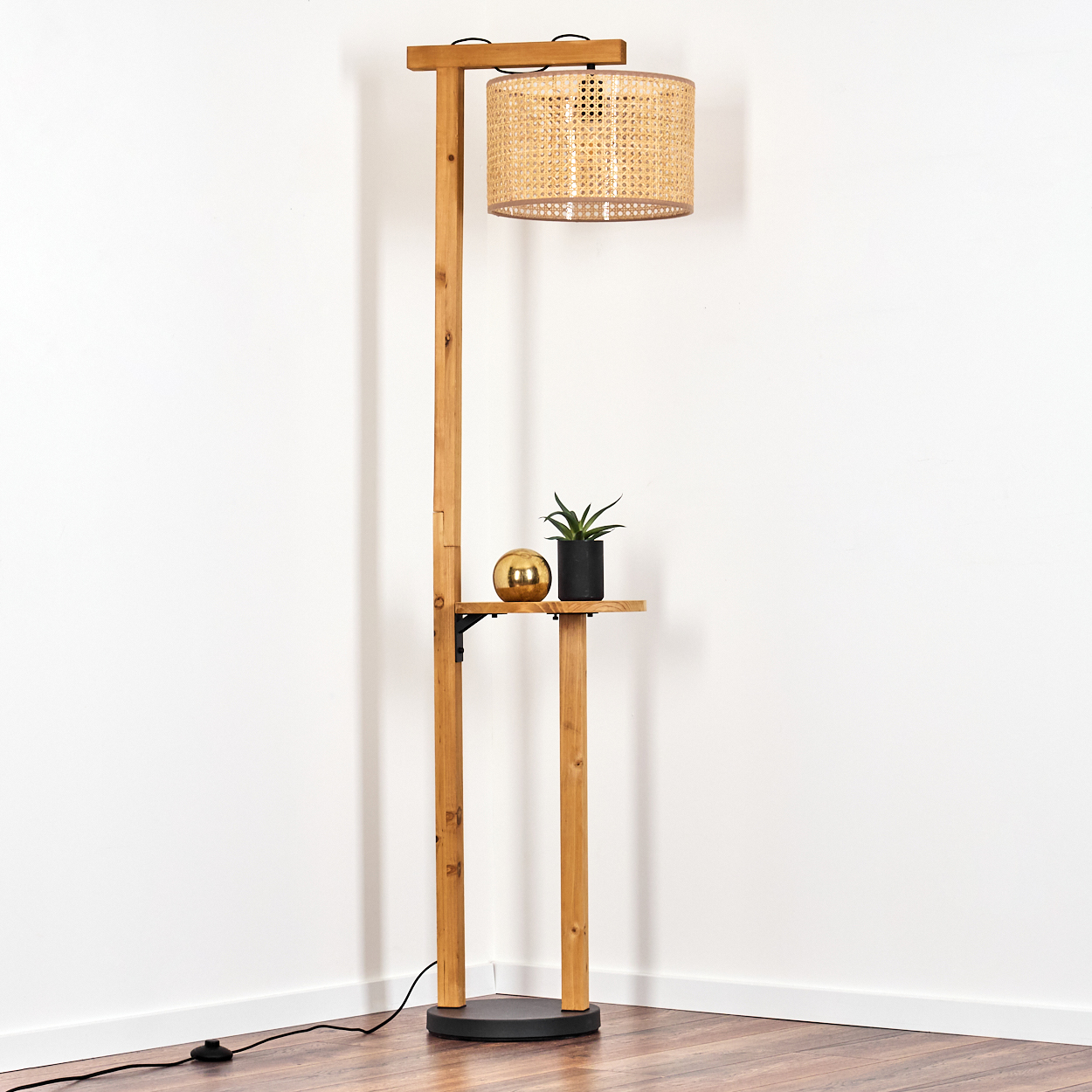 Stehlampe aus Holz mit Rattanschatten, Regal mit Pflanze und goldener Kugel, vor weißer Wand.