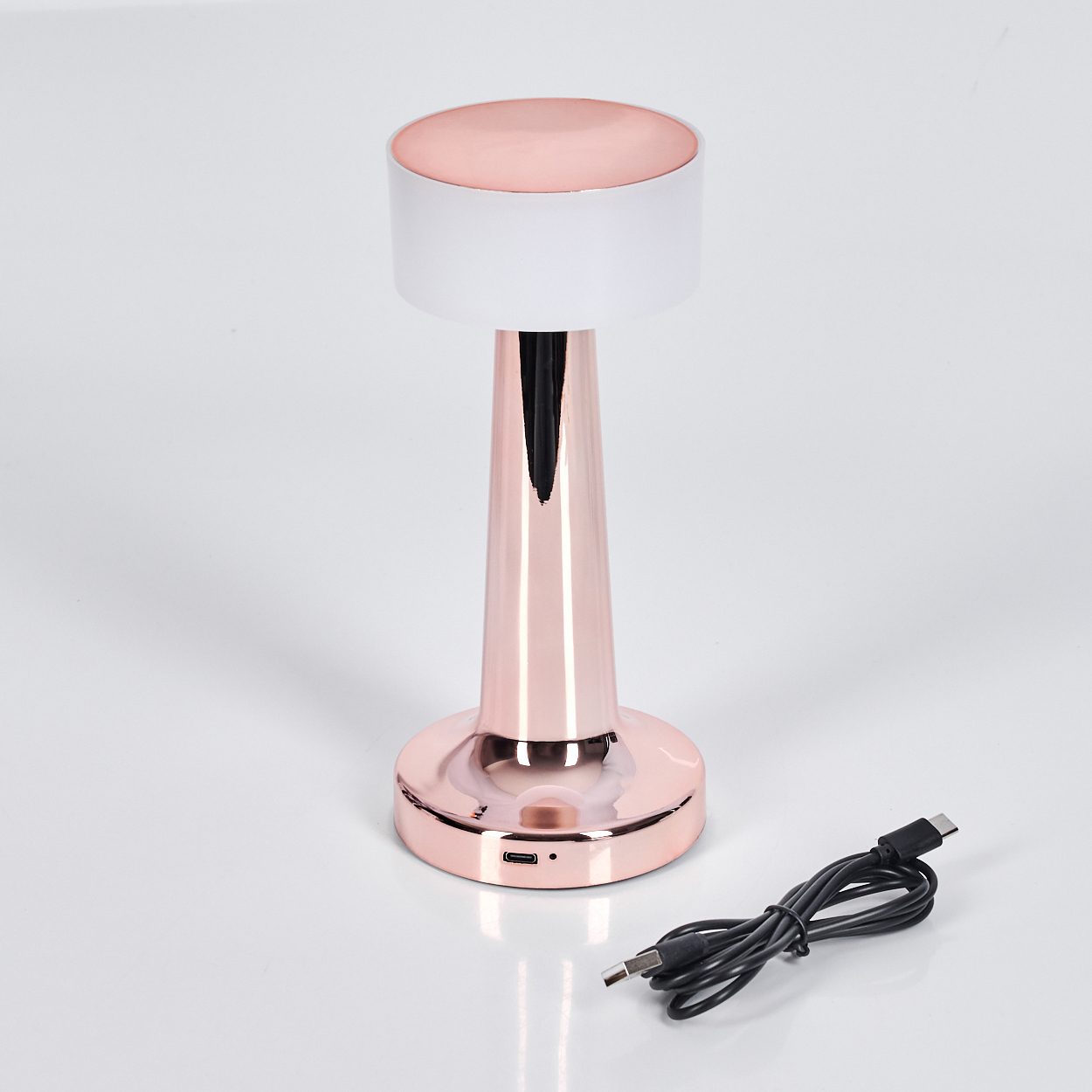 Eine roségoldene Tischlampe mit weißem Lampenschirm. Ein schwarzes USB-Kabel liegt neben der Lampe auf einer weißen Oberfläche.