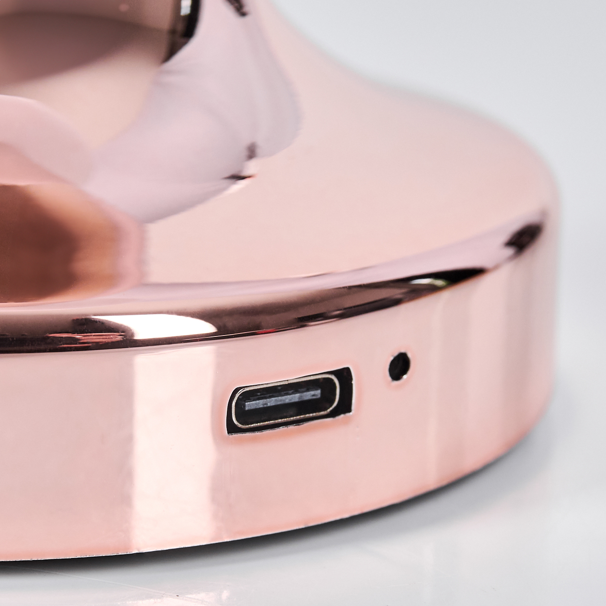 Nahaufnahme eines roségoldenen Objekts mit USB-C-Anschluss und einem kleinen Loch. Das Objekt reflektiert Licht und steht auf einer weißen Oberfläche.