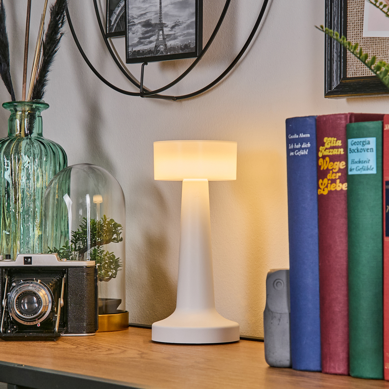 Eine weiße Lampe, Bücher, eine Kamera und Dekorationsgegenstände stehen auf einer Holzoberfläche. Ein Schwarz-Weiß-Foto hängt an der Wand.