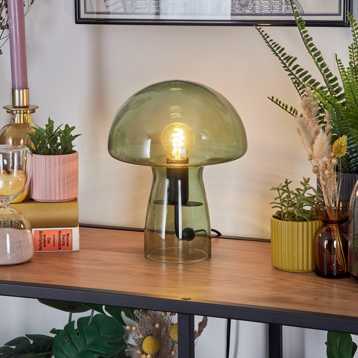 Eine grüne Pilzlampe auf einem Holzregal, umgeben von Pflanzen, einer Kerze und einem gerahmten Bild. Die Lampe ist an.