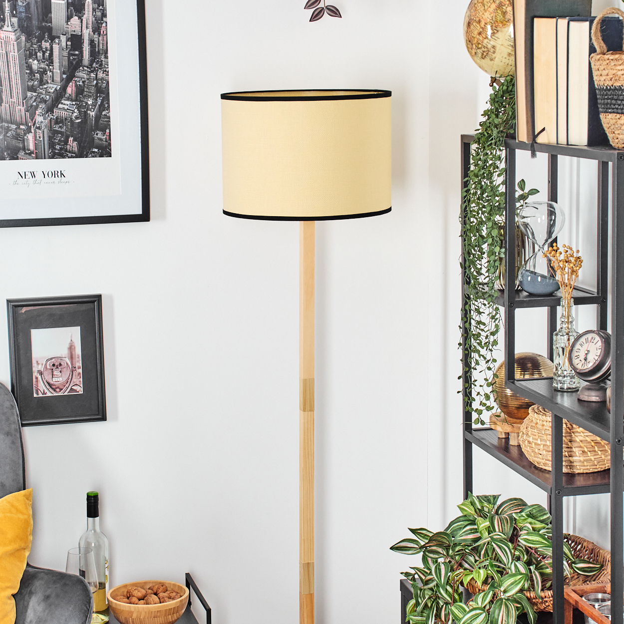 Ein Rauminterieur mit Stehlampe, Regal mit Deko und gerahmten Bildern. Die Lampe hat einen cremefarbenen Schirm und schwarzen Rand.