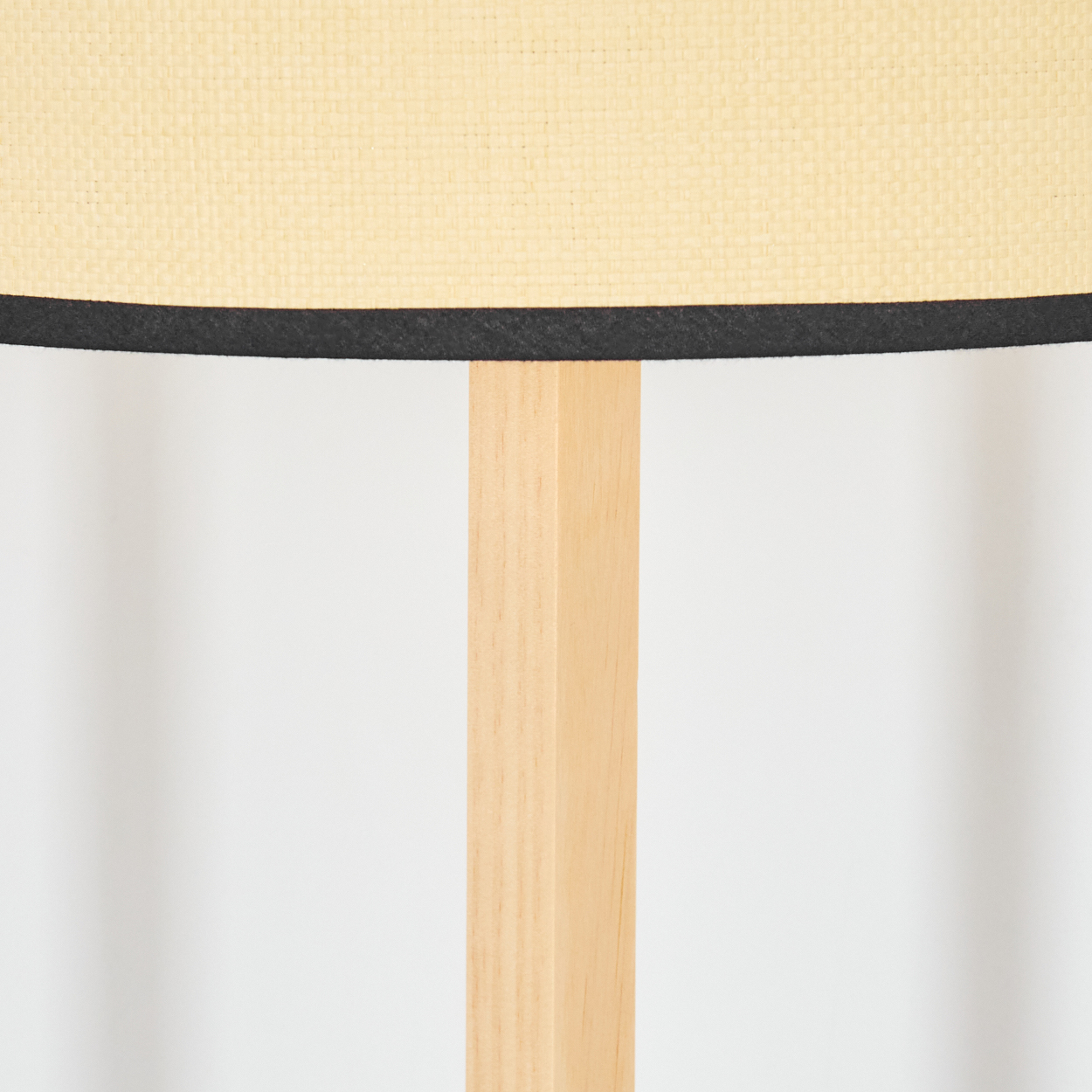 Eine Lampe mit hellbeigem Schirm, schwarzem Besatz und hellem Holzständer. Der Hintergrund ist weiß.