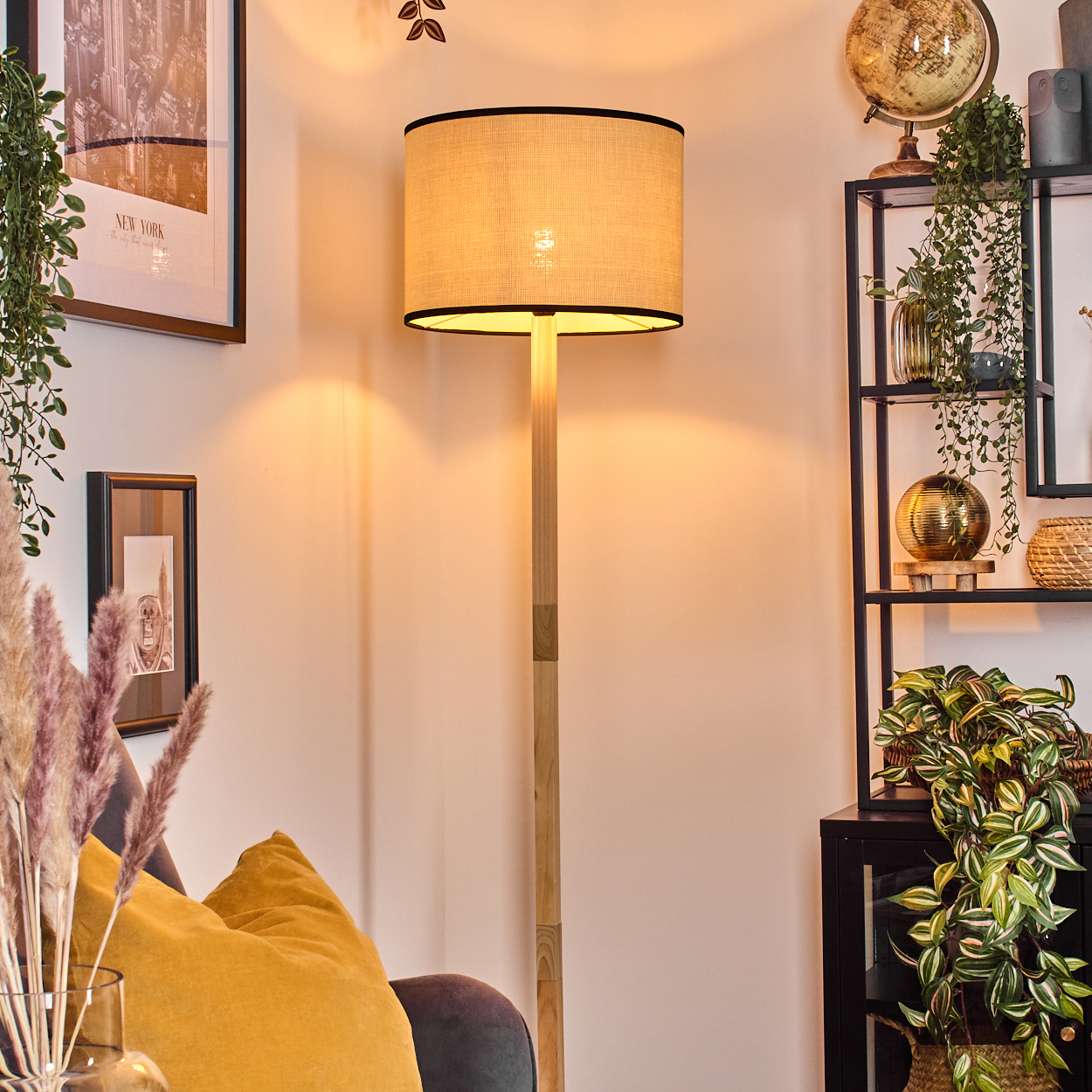 Eine beleuchtete Stehlampe steht in der Nähe eines Regals mit Pflanzen und einem Globus. Ein gelbes Kissen und gerahmte Kunstwerke befinden sich im Vordergrund.
