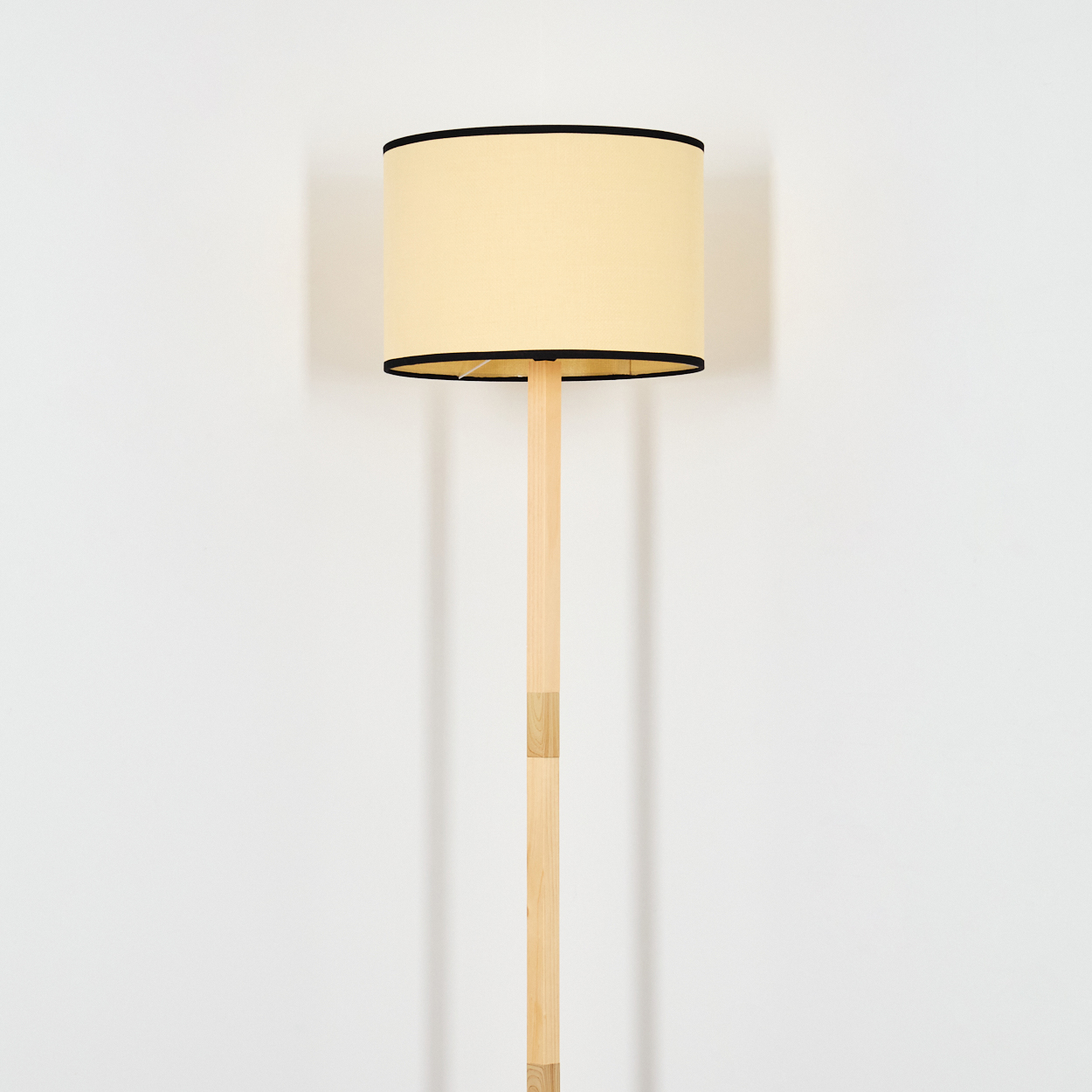 Eine Stehlampe aus Holz mit cremefarbenem Lampenschirm und schwarzem Rand. Die Lampe steht vor einer weißen Wand und wirft einen Schatten.