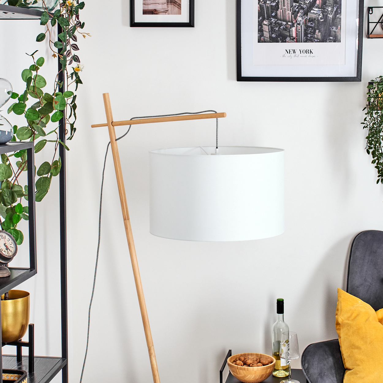 Eine weiße Lampe mit Holzrahmen steht in einem Raum. Links ein Regal mit Pflanzen. Ein New York-Bild an der Wand.