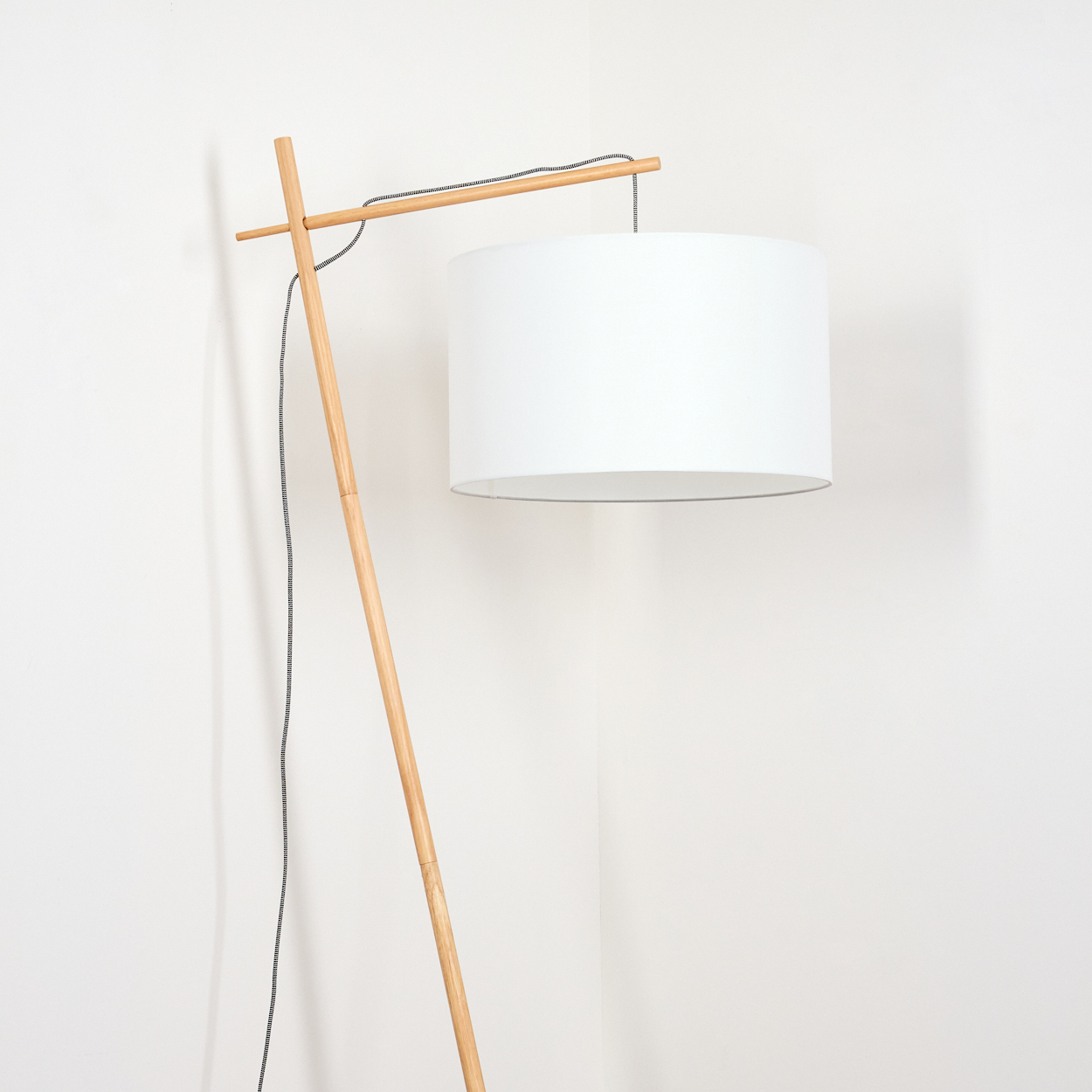 Eine moderne Stehlampe mit weißem Lampenschirm und Holzrahmen steht vor einer weißen Wand. Ein schwarz-weißes Kabel ist sichtbar.