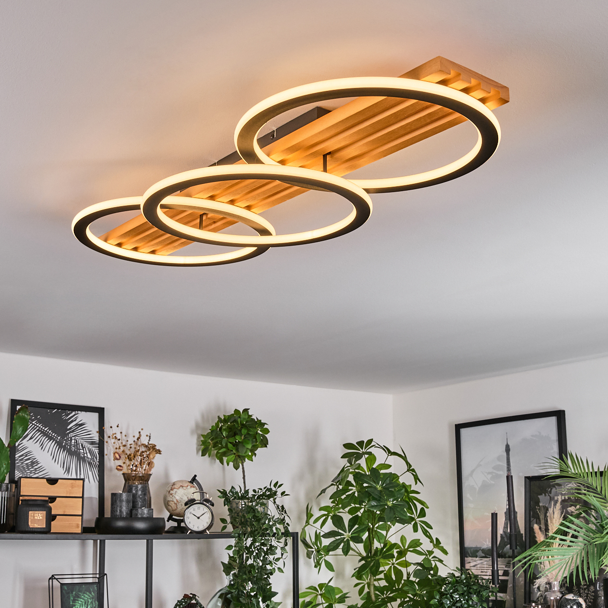 Eine moderne Deckenleuchte mit drei kreisförmigen Lichtern und einem Holzsockel. Pflanzen und Dekorationen sind unten sichtbar.