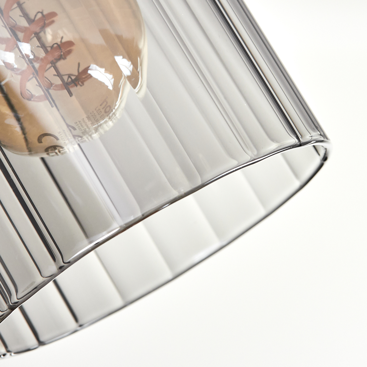 Nahaufnahme eines Lampenschirms aus Glas mit vertikalen Rillen. Im Inneren ist eine Glühbirne zu sehen. Der Hintergrund ist weiß.