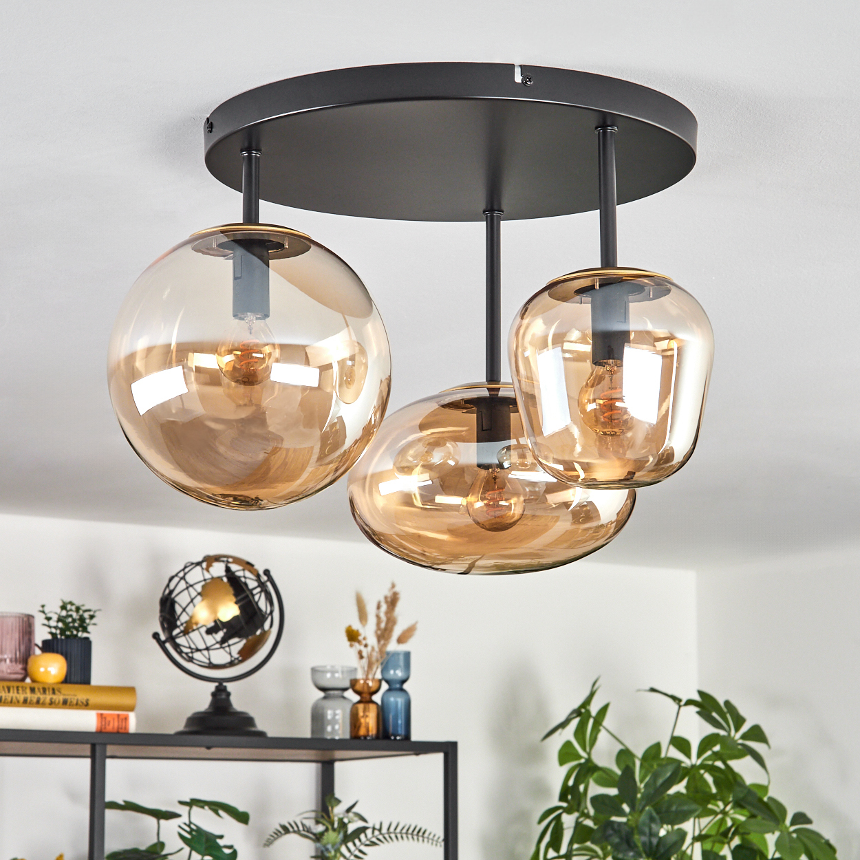 Eine Deckenleuchte mit drei bernsteinfarbenen Glasschirmen hängt über einem Regal mit Dekoration. Ein Globus und Pflanzen sind ebenfalls zu sehen.