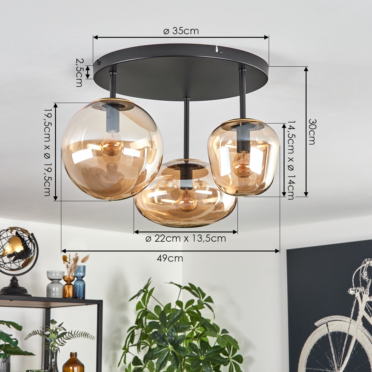 Eine Deckenlampe mit drei bernsteinfarbenen Glasschirmen. Abmessungen sind angegeben. Im Vordergrund befinden sich ein Regal mit Dekorationen und eine Pflanze.